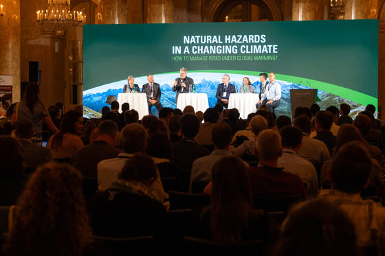 View on the stage of the Interpraevent 2024 stage for the panel discussion with the 8 panellists and moderator in front of high tables and in the background the screen with the titel of the conference